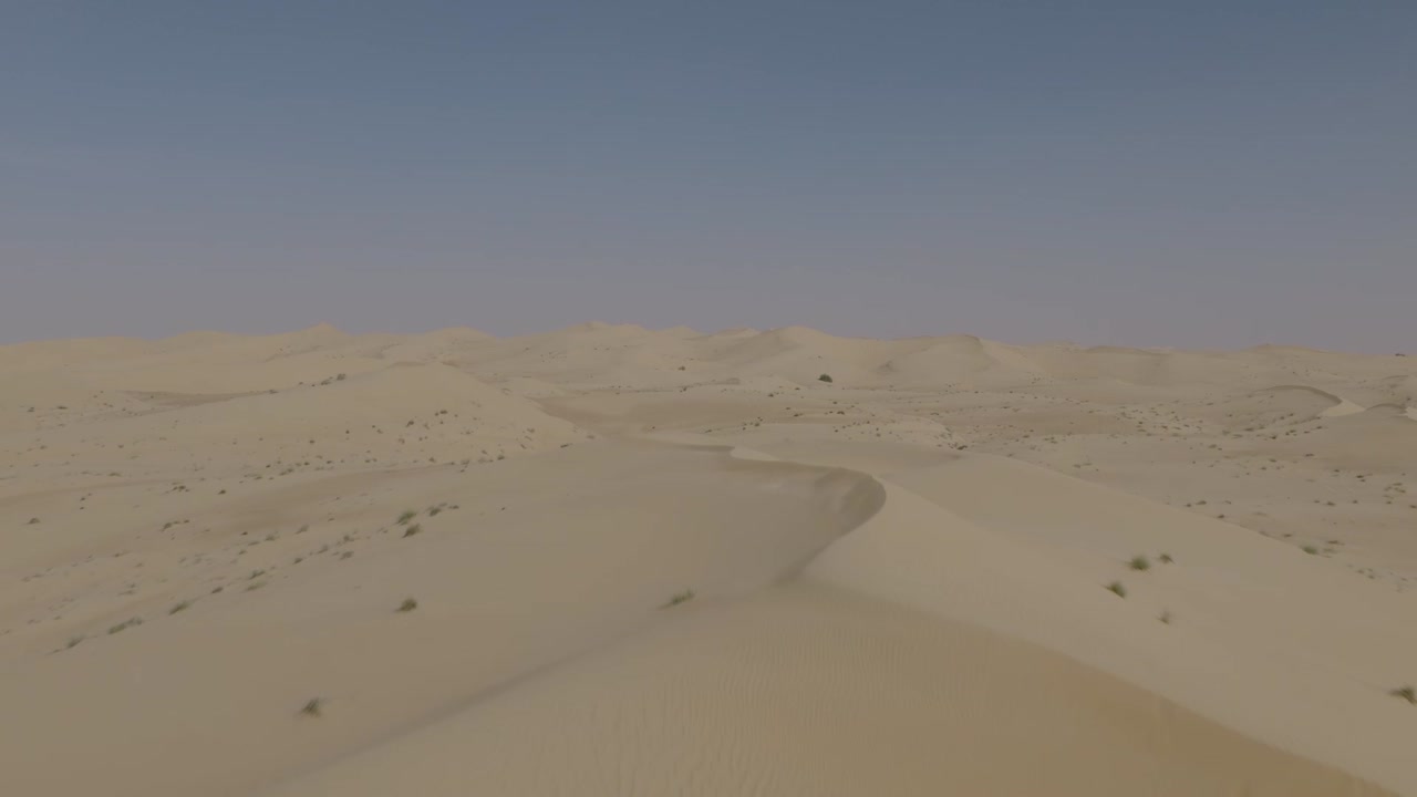 Una ripresa con drone pulita e stabile che avanza e sale sopra dune di sabbia morbide e ondulate. Il vasto paesaggio desertico incontra un orizzonte limpido, offrendo un senso di pace e di immensa vastità