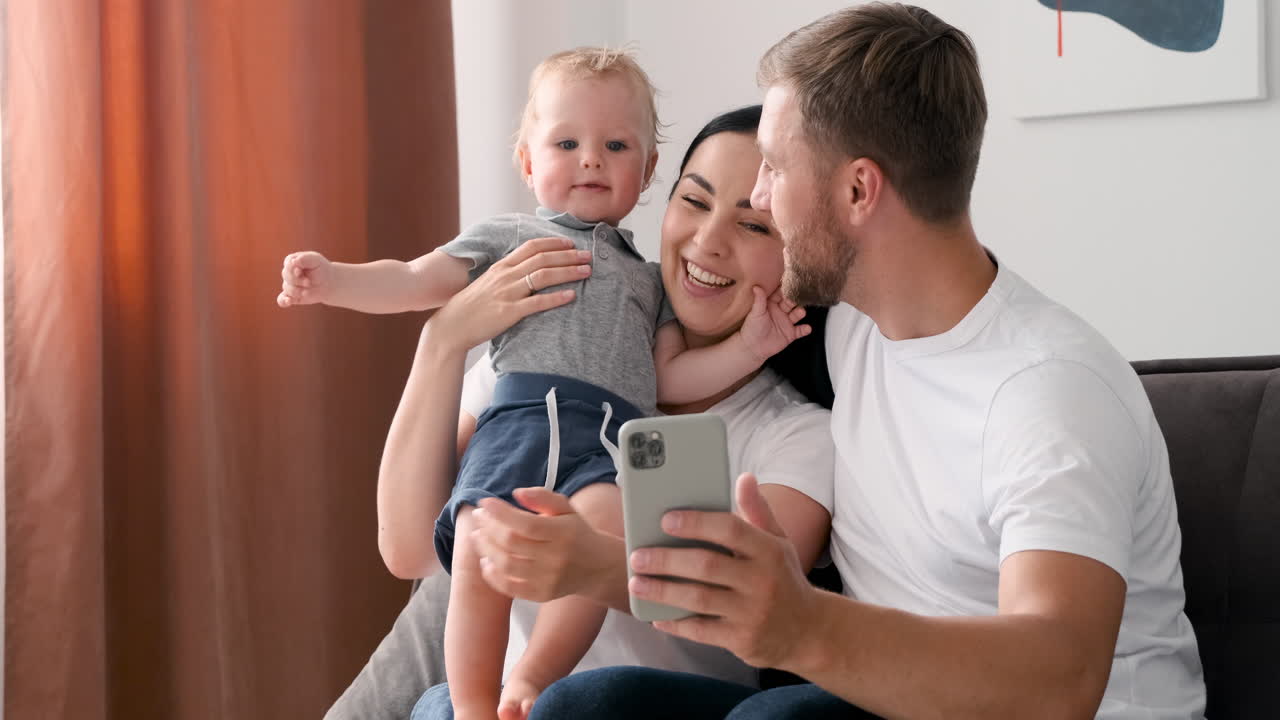 Loving Parents Sitting On Sofa In Living Room With Their Adorable Little Boy Having A Video Call On Smartphone