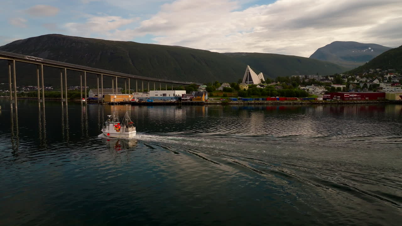 Drone past fishing boat headed toward Tromso bridge with view of Arctic cathedral