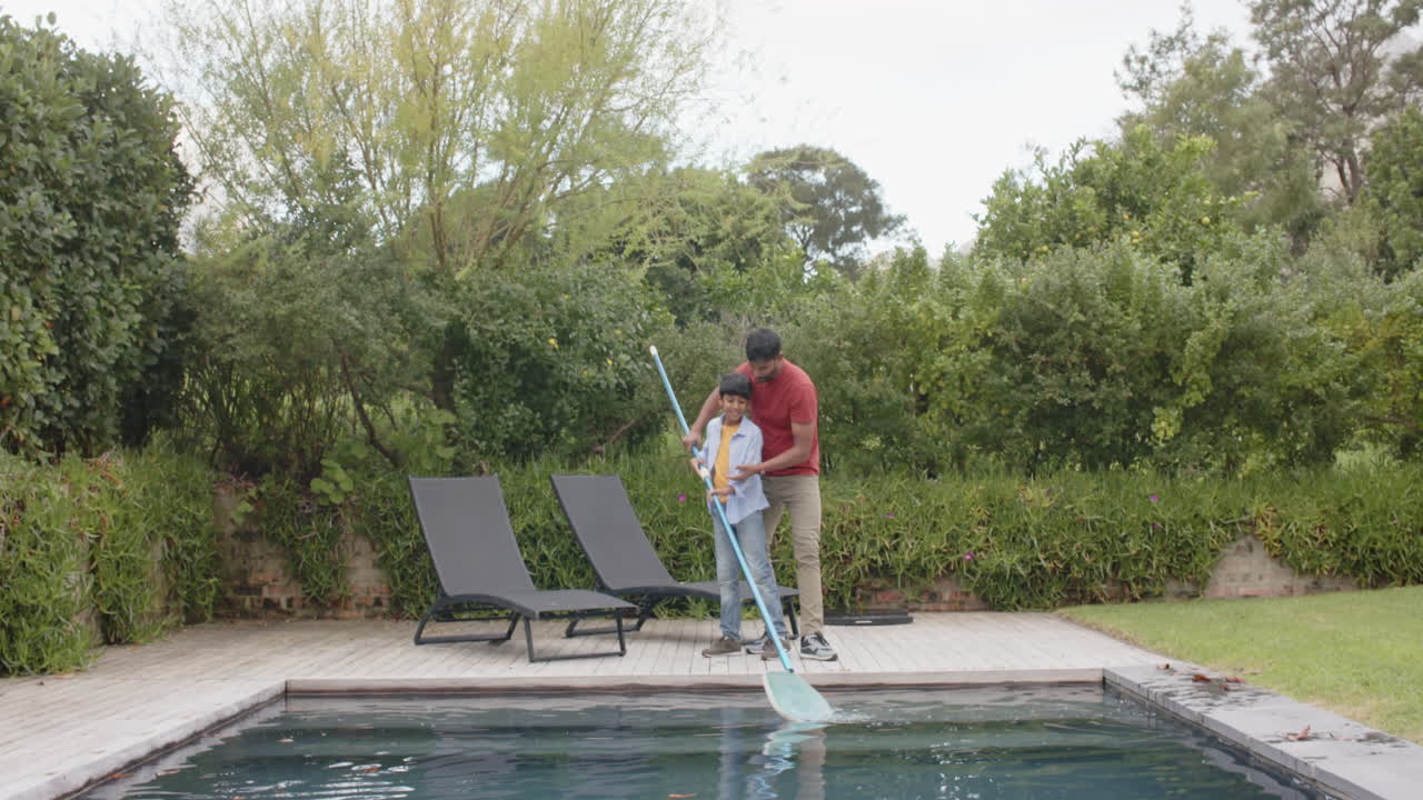 Cleaning swimming pool, young boy using pool skimmer with Indian father supervising