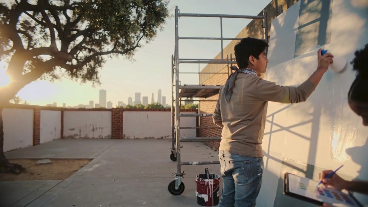 Painting man rolling paint on rooftop wall at start, woman writing on tablet, planning mural