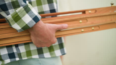 un primer plano de un hombre con una camisa a cuadros verde y azul sosteniendo un conjunto de listones de madera, revelando el grano de madera y los tornillos de oro. fondo borroso de una pared blanca y césped verde