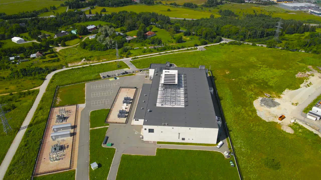 Aerial drone footage orbiting around a large and modern white and gray colored data centre in sunny countryside that has a power grid substation next to it and cooling system fans on the roof spinning