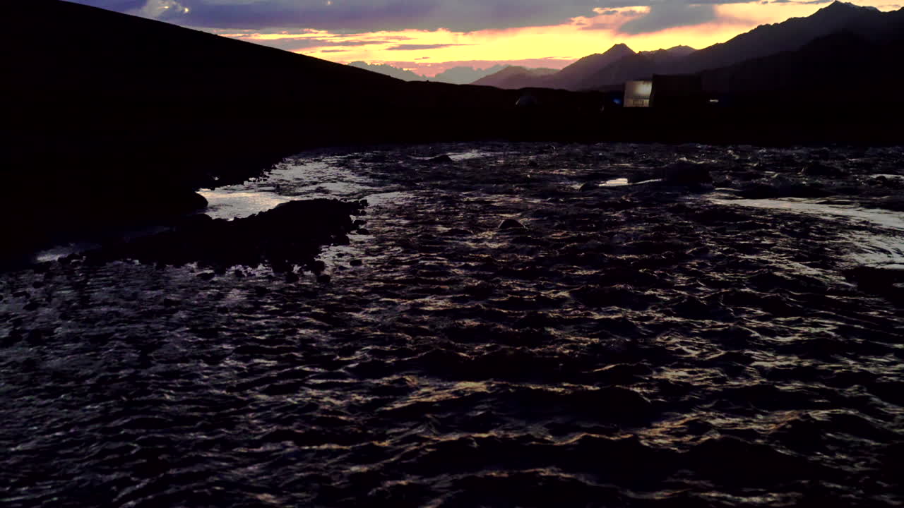 강 너머에서 텐트의 실루엣이 있는 산의 경치, 파란색과 주황색 일몰의 베이스 캠프까지 틸트 업 샷