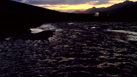 Tiro-Inclinado-Desde-Un-Río,-A-Un-Paisaje-Montañoso-Con-Siluetas-De-Tiendas-De-Campaña,-Campamento-Base-En-Puesta-De-Sol-Azul-Y-Naranja