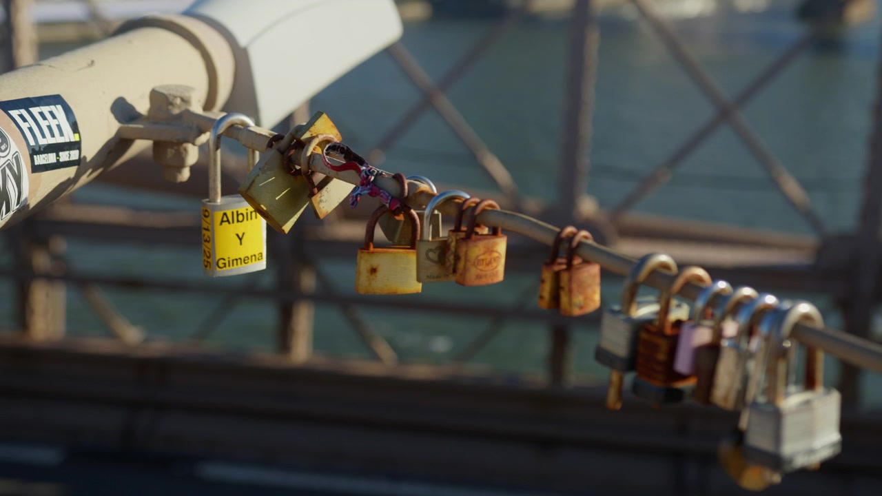 Long row of padlocks, several engraved with the names of couples, runs along a bridge cable with the river and mesh soft behind in warm afternoon light.