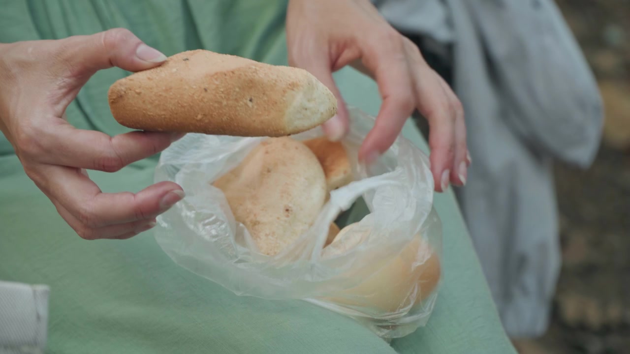 Primo piano di una donna che apre e maneggia sacchetti di plastica contenenti pane. L'attenzione rimane sulle sue mani mentre la donna scioglie il sacchetto rivelando prodotti da forno all'interno. L'illuminazione soffusa evidenzia i dettagli della texture.