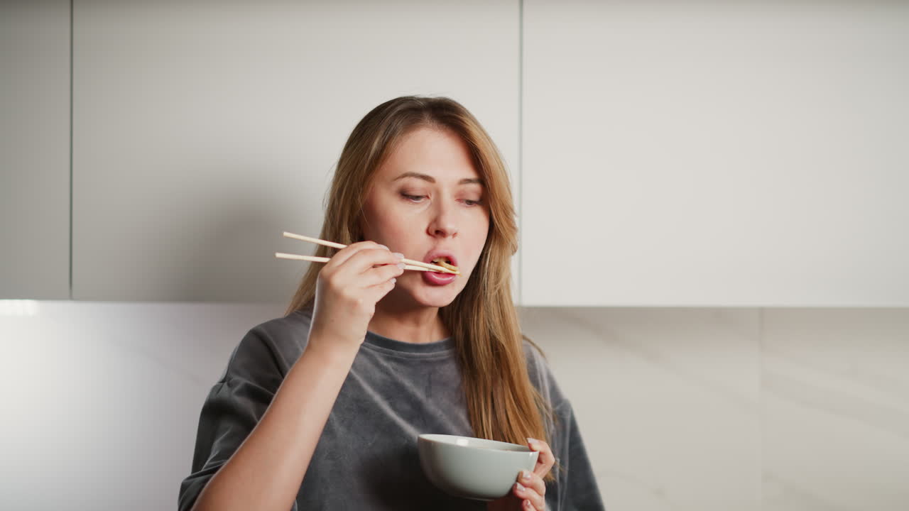 Young woman with long flowing hair eating noodles with chopsticks while standing in bright modern kitchen, savoring quick homemade meal in casual atmosphere