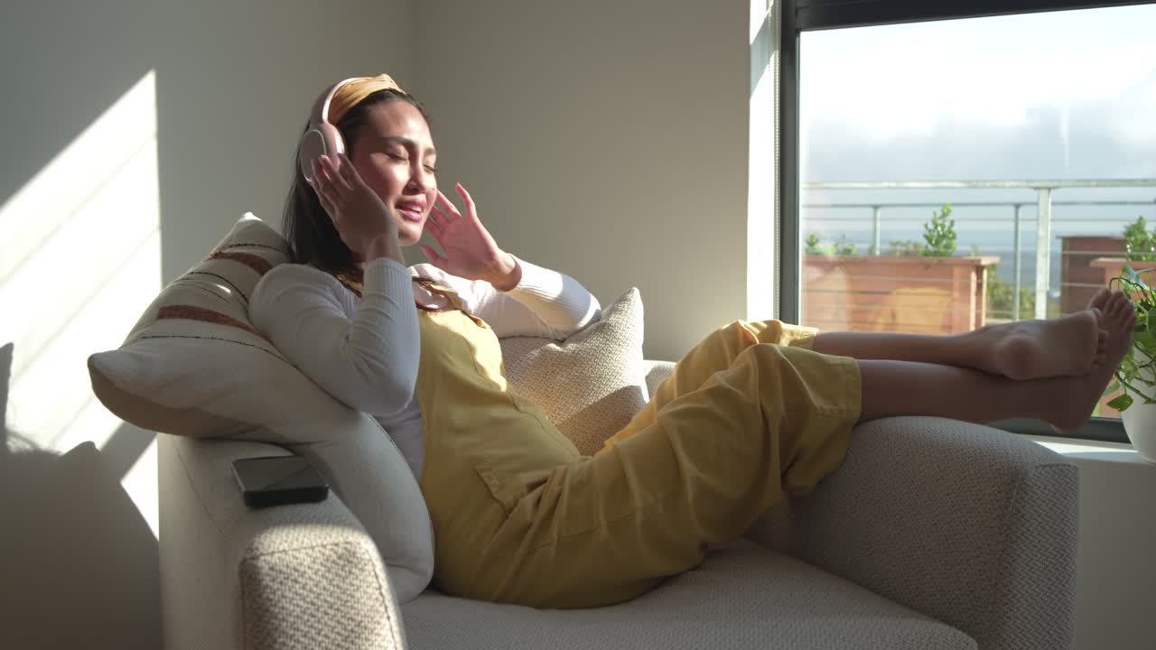 Donna seduta in poltrona, illuminata dalla luce del sole, che sistema le cuffie e scorre il telefono per scegliere la musica