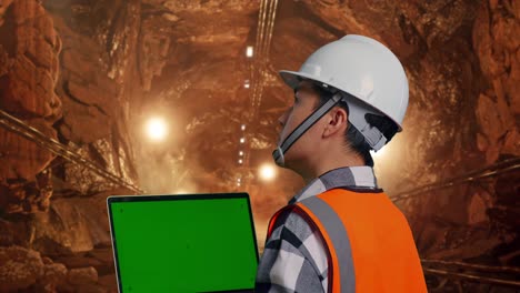 close up back view of asian male engineer with safety helmet working on a green screen laptop and looking around while standing in underground mine tunnel