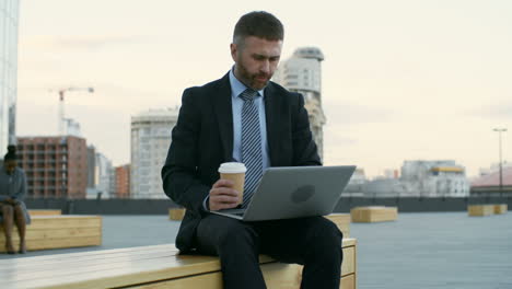 businessman sitting outdoors on wooden bench with laptop on his knees, typing on keyboard and drinking coffee