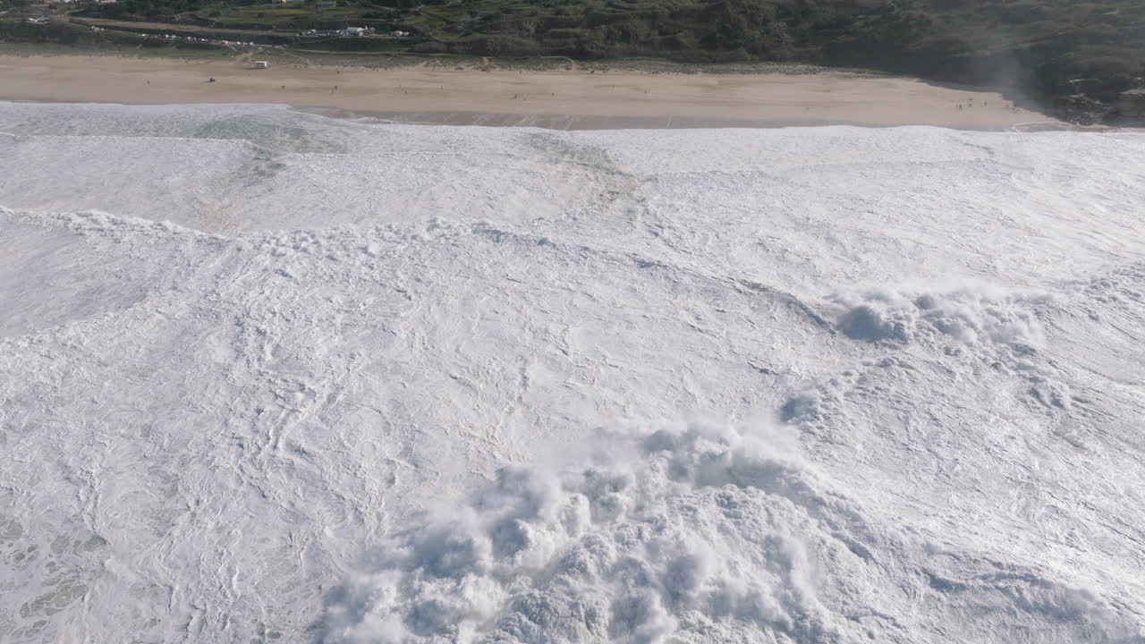Aerial drone shot of huge waves coming into shore on a day with giant waves in Nazaré, Portugal, Europe. Flying towards beach and crowd watching
