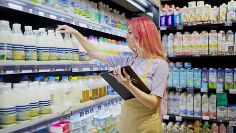 Confident-girl-with-bright-hair-in-an-apron-holds-a-tablet-in-her-hands-and-takes-inventory-using-a-tablet-in-a-supermarket