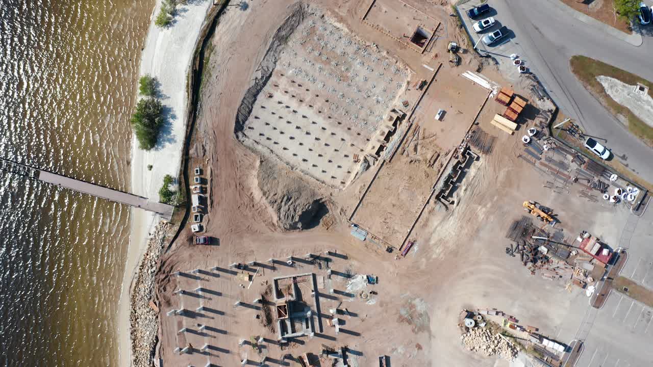 Construction site by the coast featuring concrete foundations and structural columns adjacent to a parking area with vehicles, next to a body of water and a small sandy shoreline