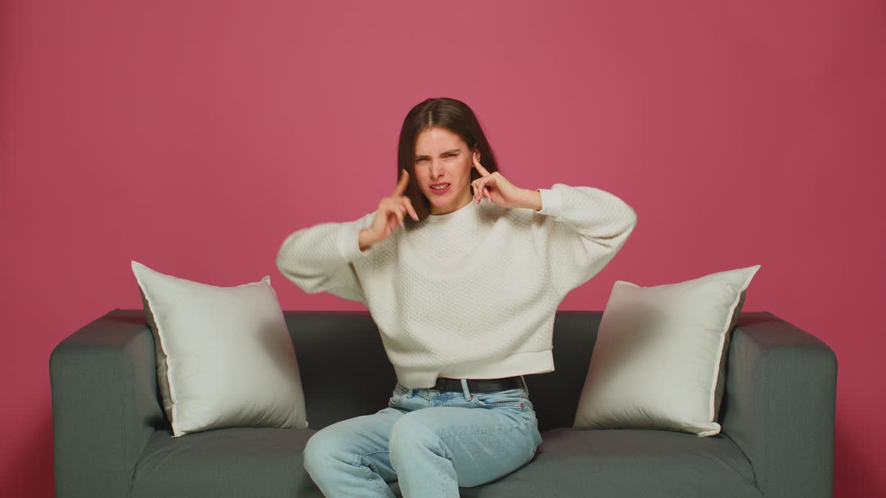 짜증나는 어린 소녀는 귀에 손가락을 넣고 듣지 않고, 시끄러운 이웃을 듣기를 거부하고, 집에서 소파에 앉습니다.