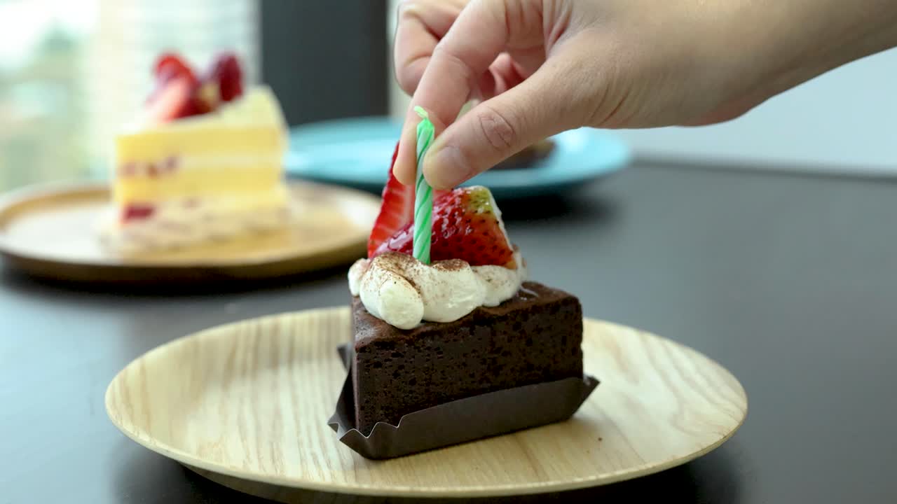 Hand inserts green candle into chocolate cake slice with strawberry, natural light, shallow focus