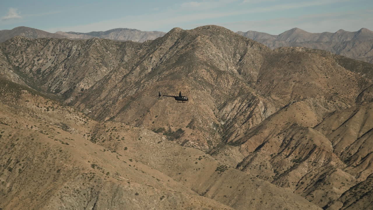 A helicopter navigates the rugged mountain terrain, showcasing the natural beauty of steep slopes and distant peaks.