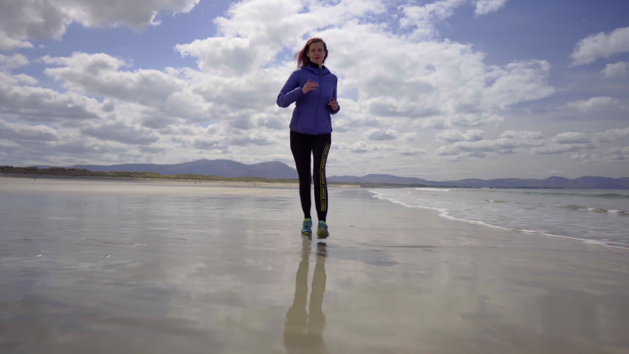 Low dolly shot of a girl running, jogging on the shore of a sandy beach with Atlantic ocean waves on a wonderful sunny day in Ireland in 4K