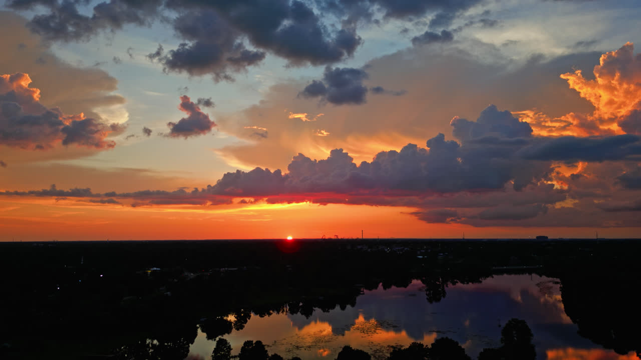 Brilliant orange and red sunset colors paint the sky over still water and forested edge, heavenly aerial backdrop background, aerial roll left