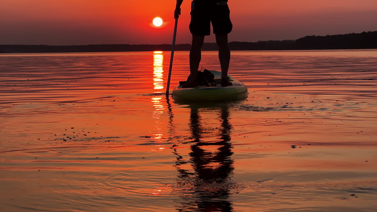Blue and yellow sup board floating by the river with man standing on. Beautiful orange sunset at backdrop.
