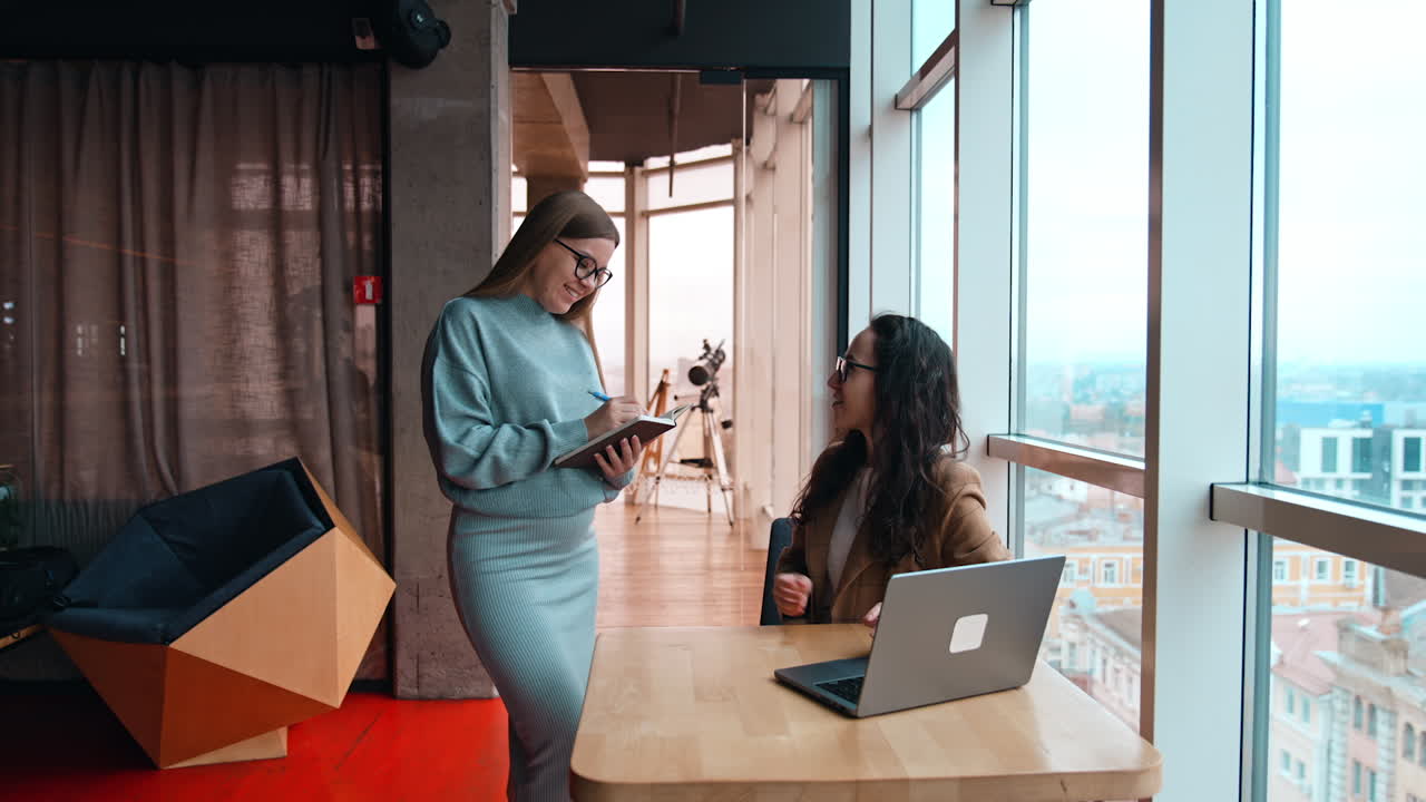 Two positive female workers communicating in the office. Blonde pregnant lady holding a notebook and taking notes. Brunette colleague sits in front of laptop.