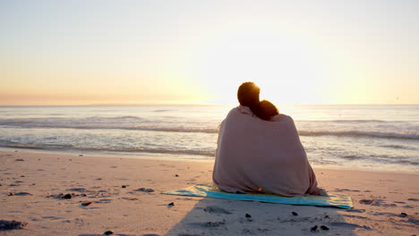 pareja biracial envuelta en una manta, viendo una puesta de sol en la playa con espacio de copia