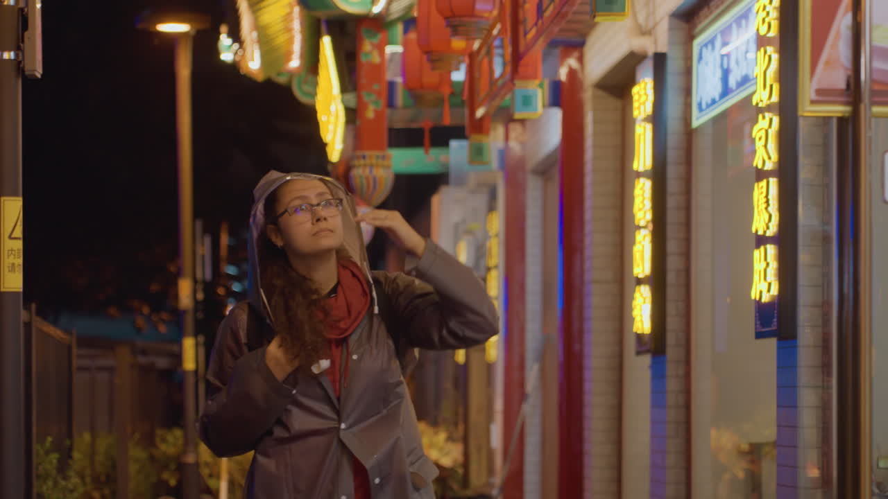 Primo piano di una donna asiatica in una strada illuminata al neon che guarda in alto, indossa cappello e occhiali, sciarpa rossa e cappotto lungo, con espressione riflessiva e sguardo dolce, turista che nota lanterne e insegne vivaci, cinematografico.