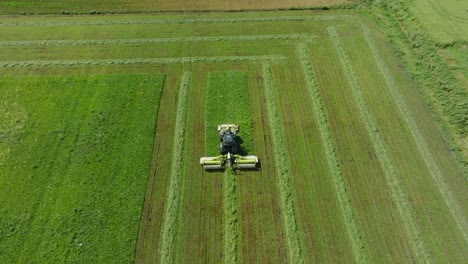 aerial establishing view of a tractor mowing a fresh green grass field, a farmer in a modern tractor preparing food for farm animals, sunny summer day, wide birdseye drone shot moving forward