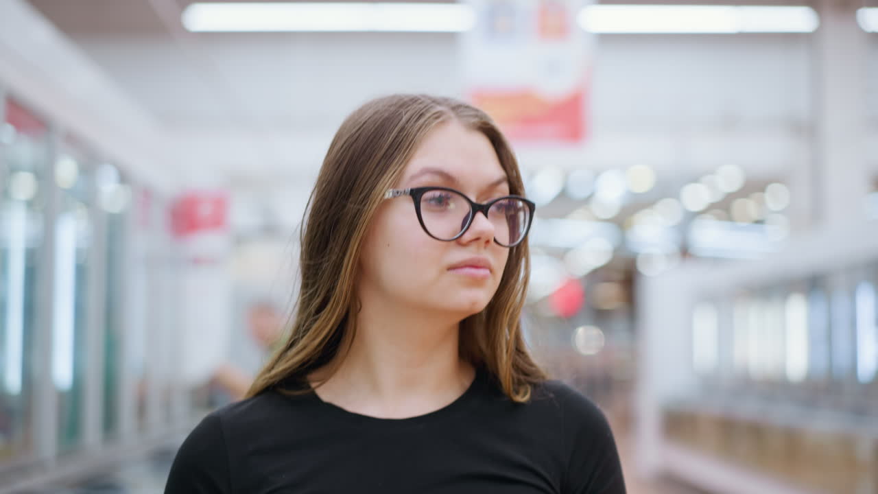 mujer de compras en la parte superior negra camina más allá del vidrio de exhibición con otro comprador en el fondo, ella sostiene bolsas de compras, mirando hacia adelante mientras se mueve a través de un ambiente de centro comercial brillante y moderno
