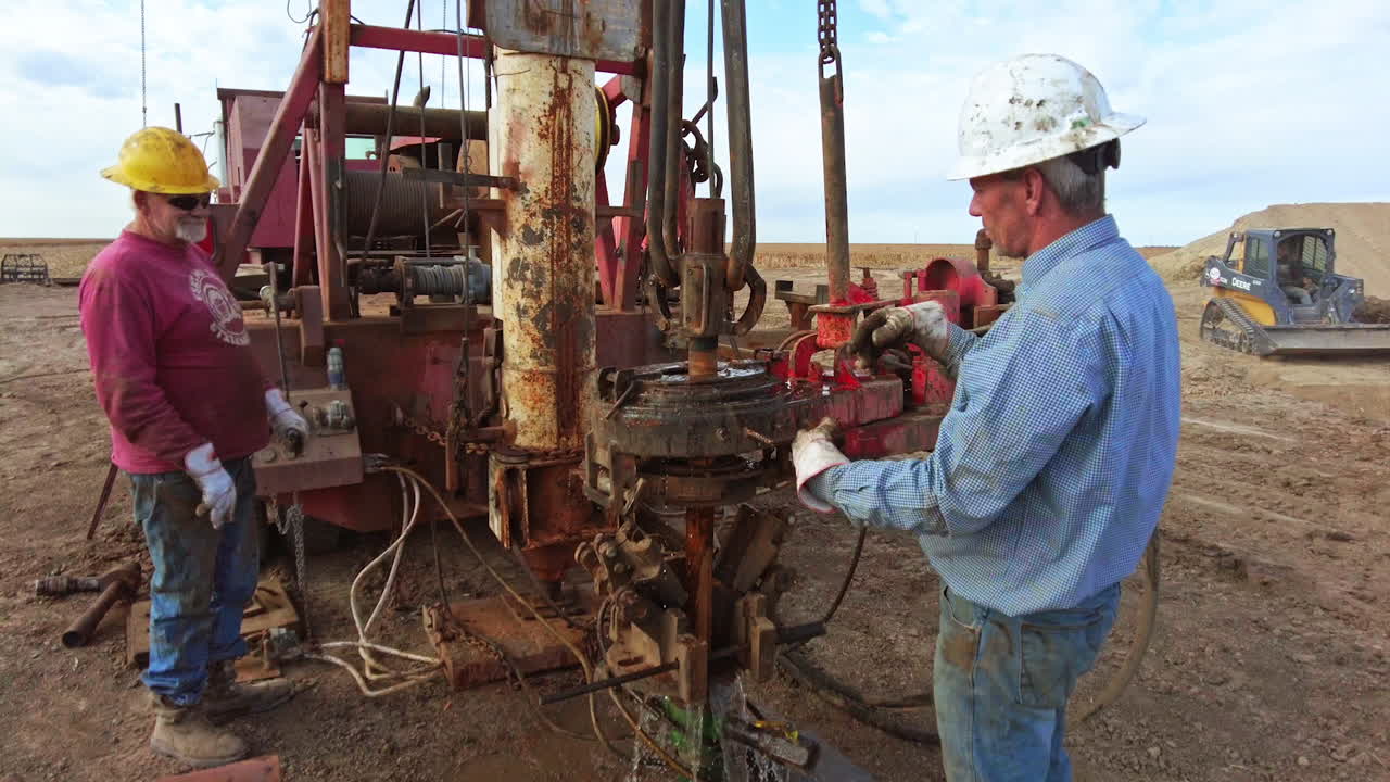 Approaching two men working at drilling equipment in the desert. A pile of metal pipes is at the truck.