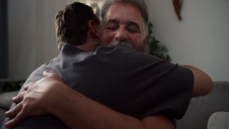 close-up-Over-the-shoulder-of-a-happy-man-with-gray-hair-and-a-lush-gray-beard-hugging-his-brunette-boyfriend-in-a-gray-T-shirt-in-a-modern-apartment-on-the-sofa-in-the-evening