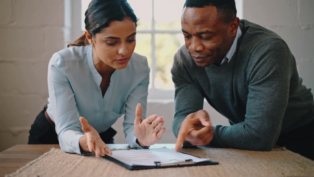 Placing hands near clipboard, woman blue-top reviewing file while man grey-top pointing office pen