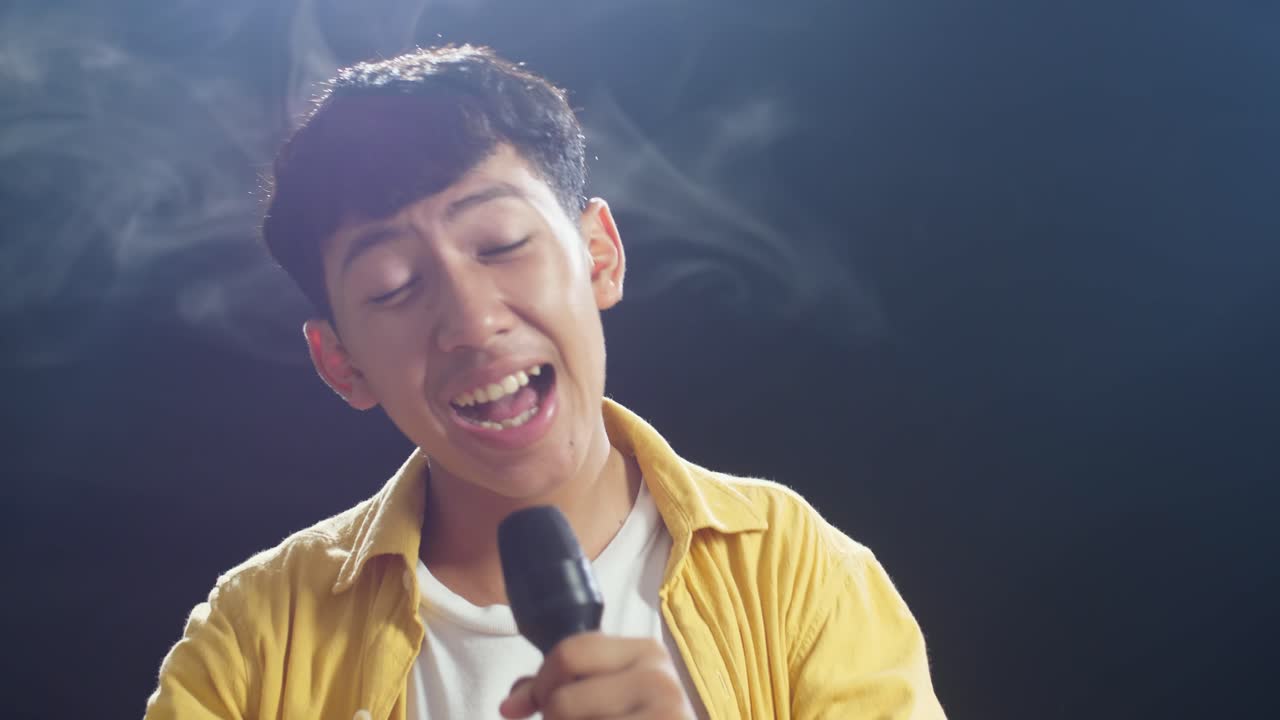 젊은 아시아 소년이 마이크를 들고 색 연기가 검은 바탕에 을 치고 있습니다.