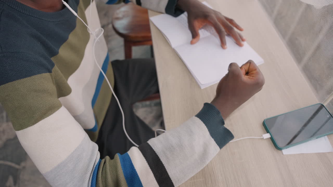 Black man writing love note at cafe table, hands holding pen over paper, striped sweater, earbuds plugged into smartphone, warm natural light, intimate closeup showing planning of romantic date
