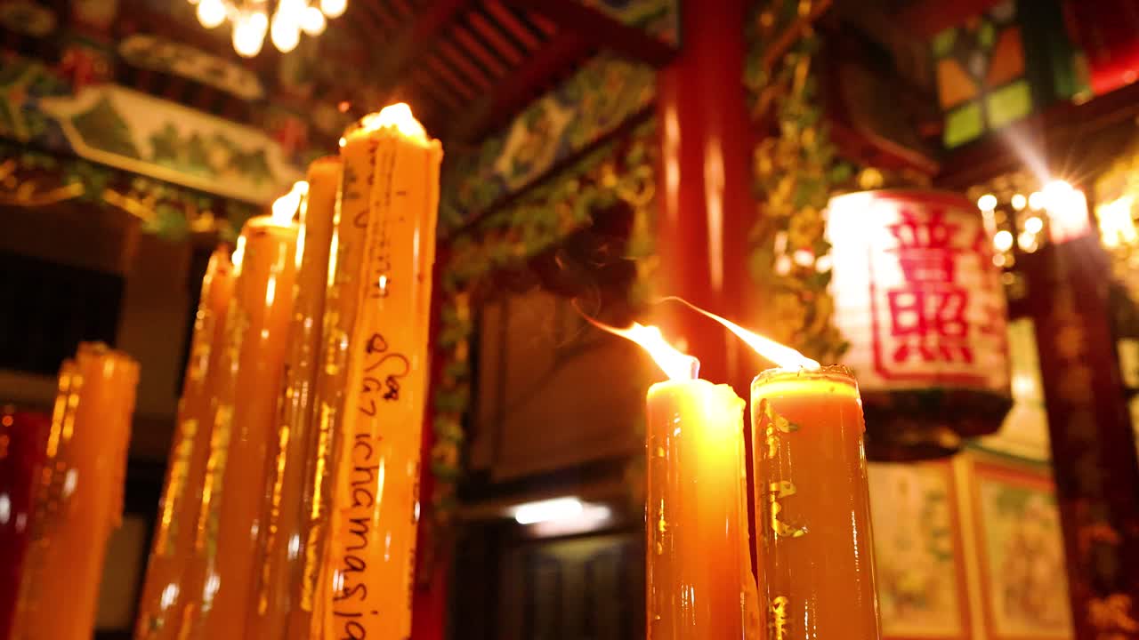 Tall candles burn in ornate Asian temple, warm lighting, low angle, slight camera movement