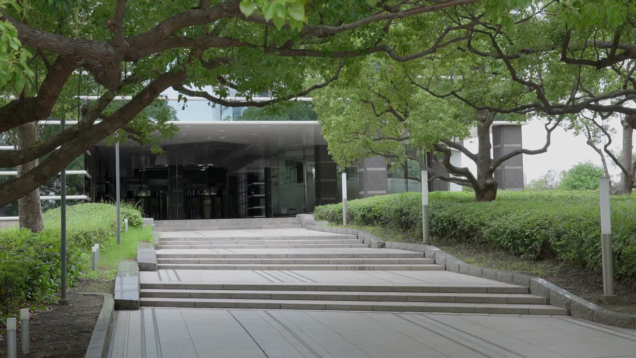 A peaceful shot of a paved walkway and stairs leading toward a modern building, surrounded by green trees and foliage