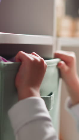 pretty blonde little girl looks inside large fabric containers on wooden rack in light children room at home close side view slow motion