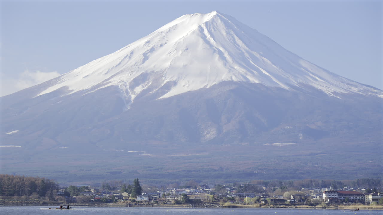 Clear skies showcase the stunning snow-capped peak of Mount Fuji, with calm waters reflecting its beauty. Nearby, a peaceful Fujikawaguchiko village nestles at the base. Kawaguchiko Lake