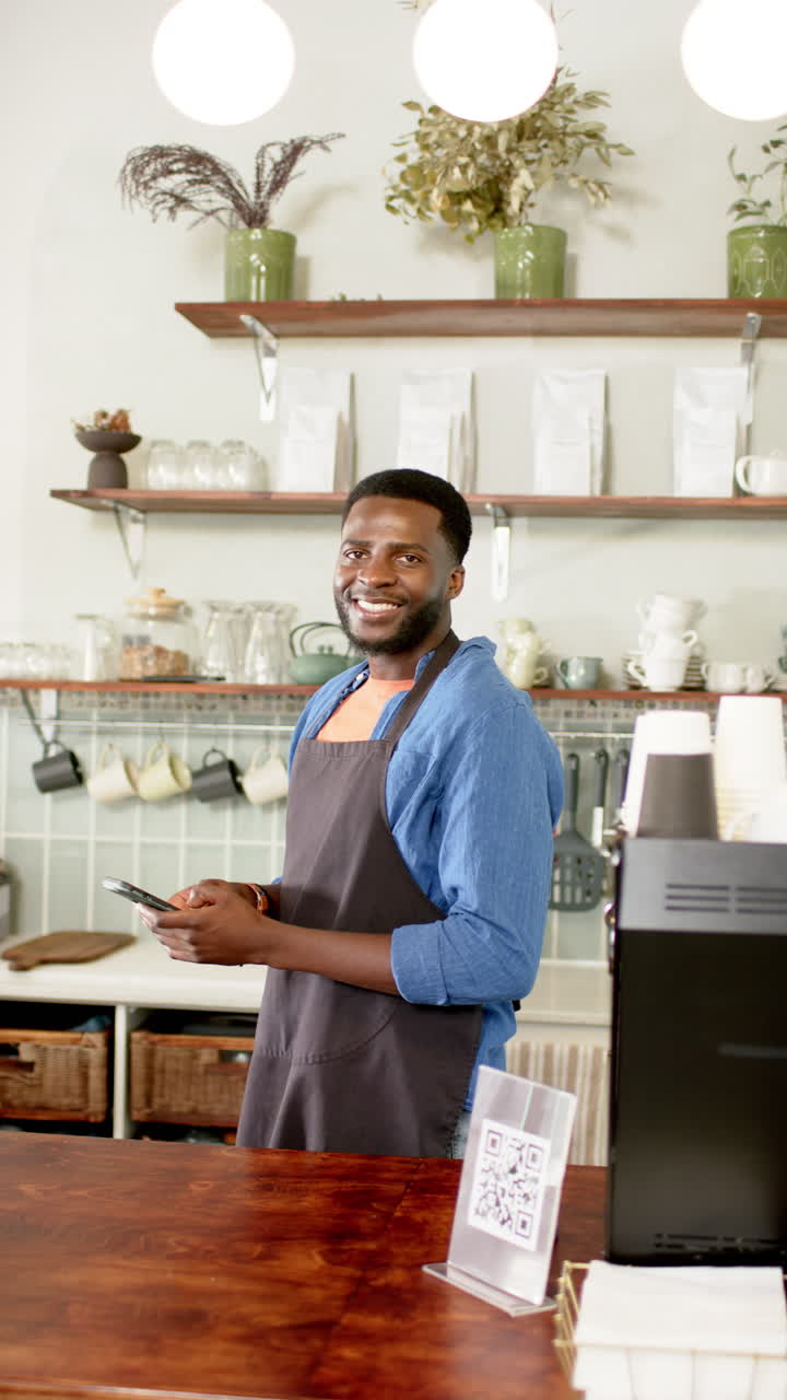 Vertical video: African American male barista smiling, holding tablet in cozy cafe