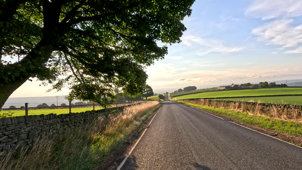 A vehicle travels along a narrow country road bordered by fields and stone walls, passing a lone tree under golden sunset light with dynamic camera movement