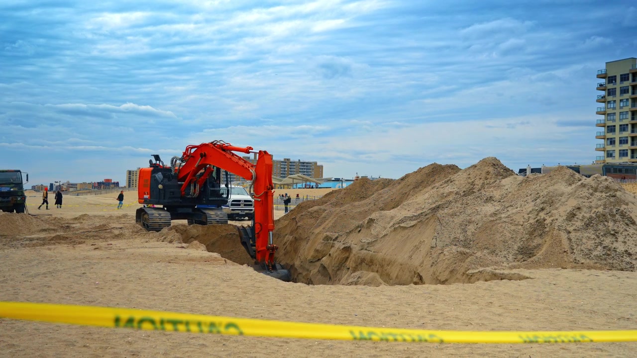 New York, USA, 1 April 2026: Orange crawler excavator digging sand on coastal beach area. Side view of heavy machinery arm digging into sand on a beach under a cloudy sky with buildings.