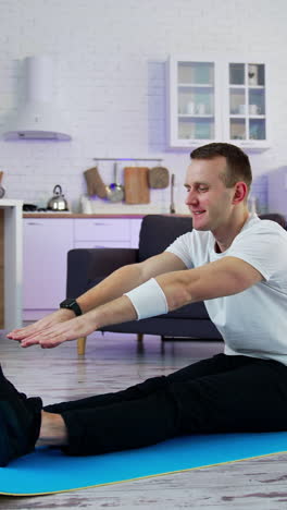 young man exercising at home. man sitting on a mat and trying to touch his feet on kitchen background. difficulties of physical training during lockdown period.. vertical video