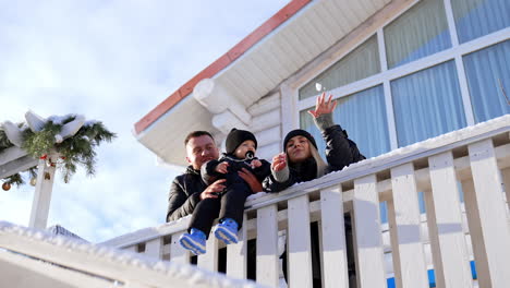 loving caucasian parents with their little son sitting on the fence stand at the porch. mother shows some snow to the baby and them throws a tiny snowball to the camera. low angle view.