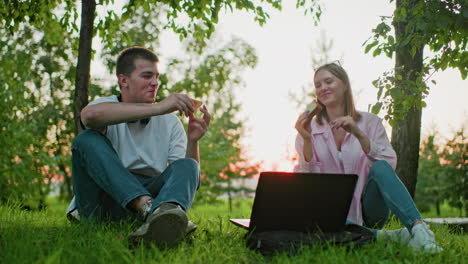 niño y niña sentados al aire libre con una computadora portátil delante, haciendo un brindis con pan en la mano, disfrutando de una comida juntos bajo la sombra de un árbol, la luz del sol filtrándose a través de las hojas en el fondo