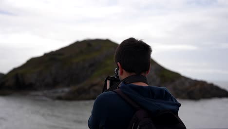 Rear-shot-of-a-man-with-a-professional-camera-capturing-photos-of-an-island-in-Bracelet-Bay,-The-Mumbles