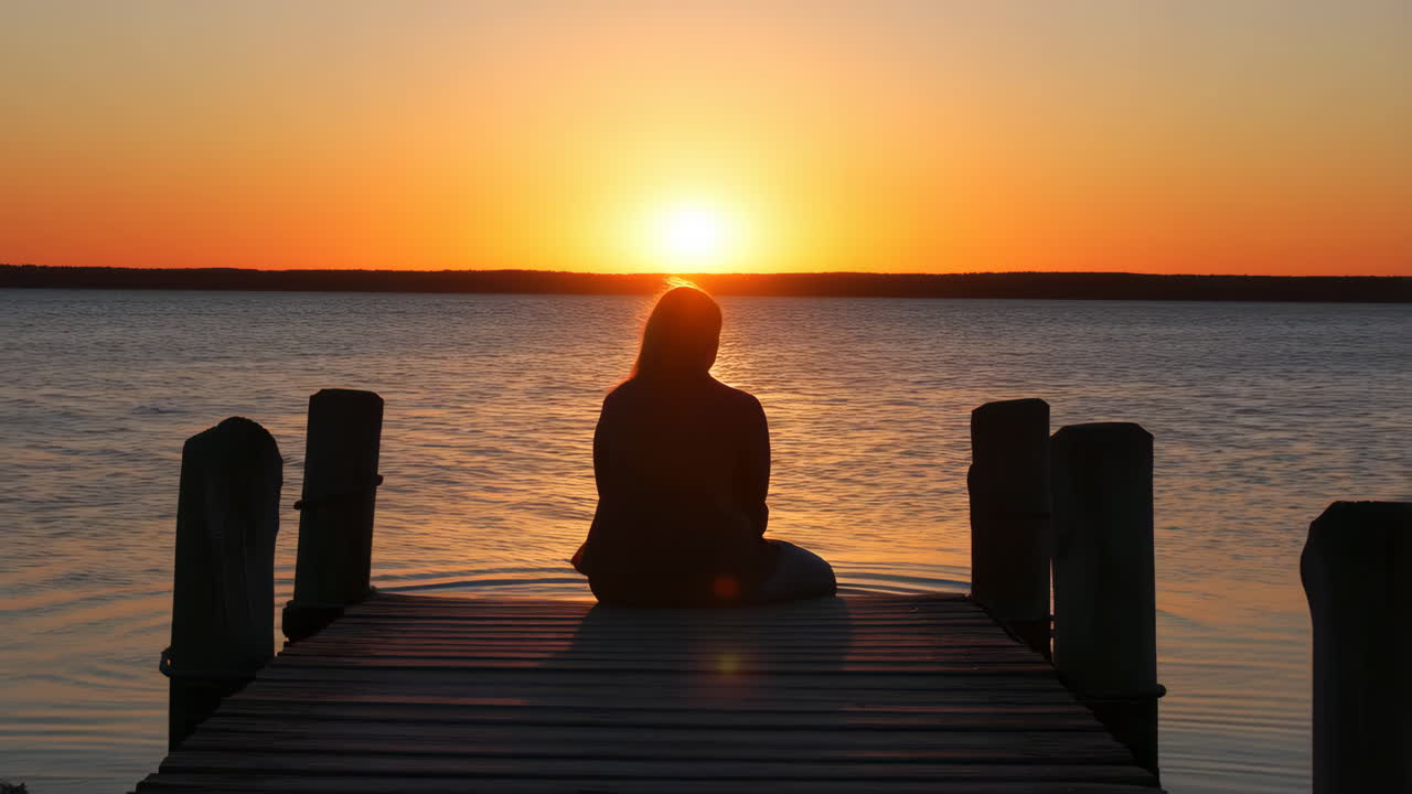 A person sits on a pier watching a vibrant sunset over water