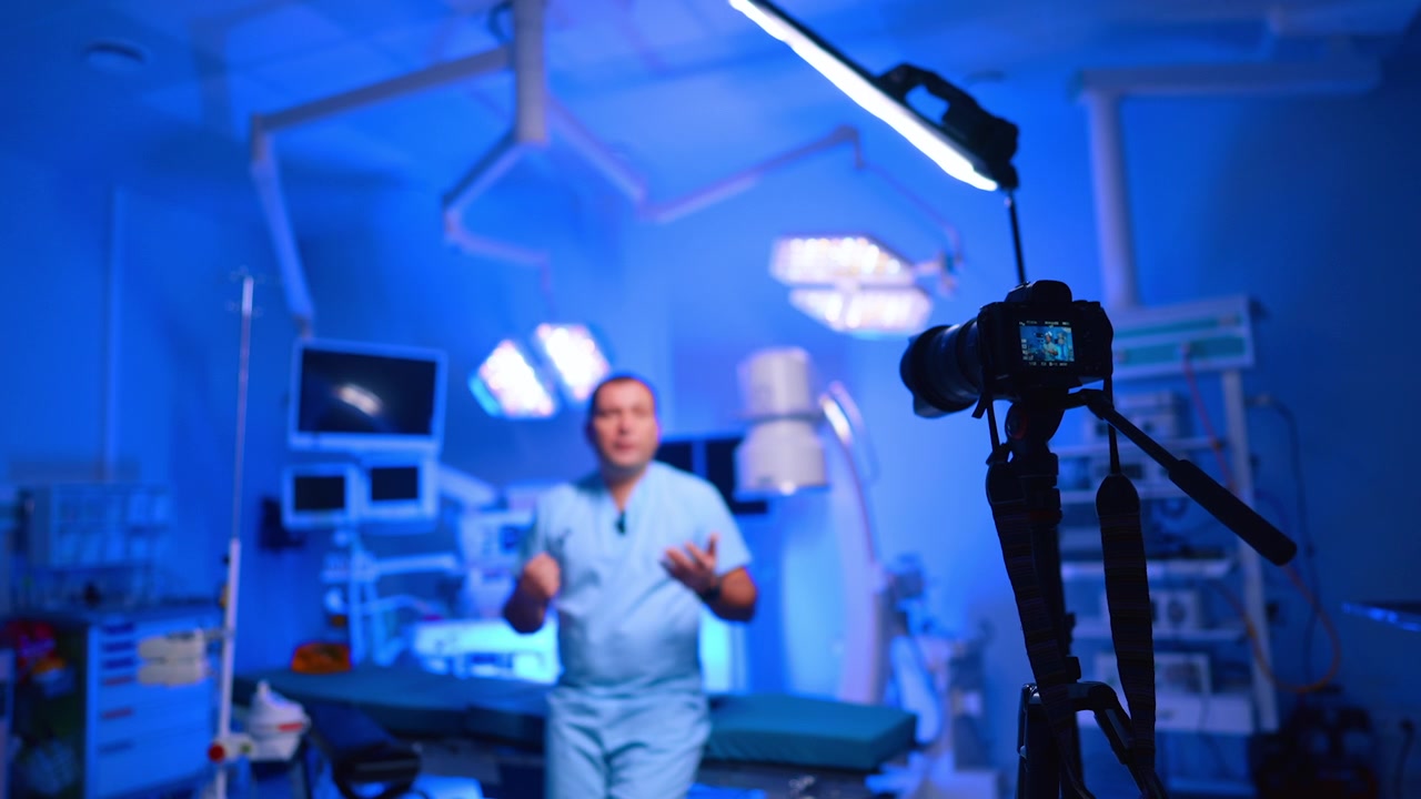 Telecamera che riprende un medico che parla in una sala operatoria illuminata di blu. Attrezzatura su treppiede con un relatore sfocato in divisa medica che gesticola sotto luci chirurgiche e monitor.