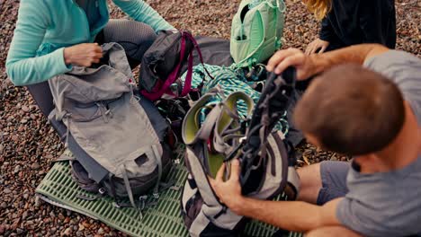 shot from above, a brunette guy in a gray t-shirt takes out special equipment and insurance for rock climbing from his backpack, along with his two girlfriends who also disassemble their backpacks and take out a rope before starting their rock climbing