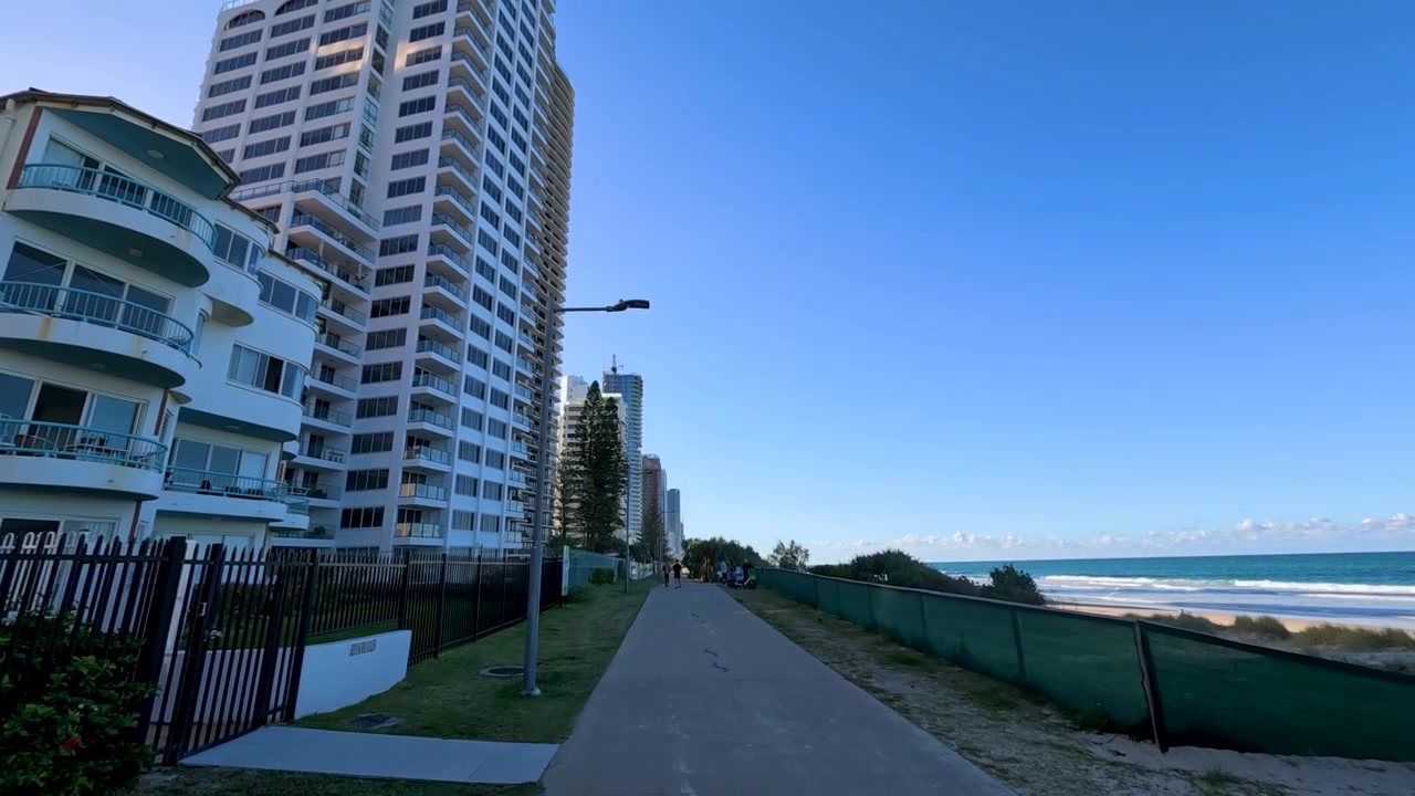 Tomada panorâmica em grande angular em movimento ao longo de um caminho costeiro ensolarado ao lado de edifícios altos em Surfers Paradise.