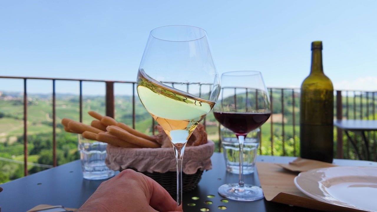Una mano fa roteare vino bianco al rallentatore contro uno sfondo panoramico della campagna italiana
